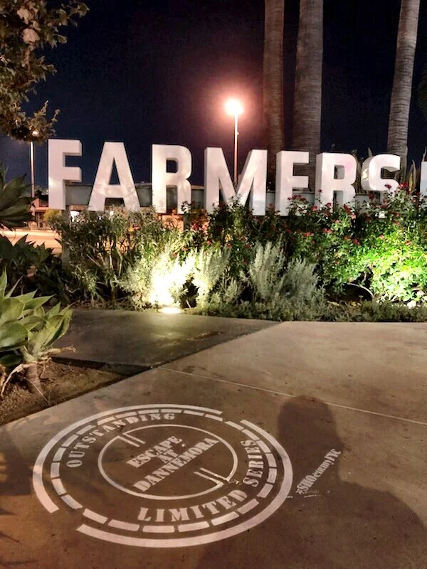 The pavement outside the Farmers Market in Los Angeles. The stencil is a manhole cover, which is how the inmates get out in Escape at Dannemora, with the category line around the rim. It cost a fraction of one billboard